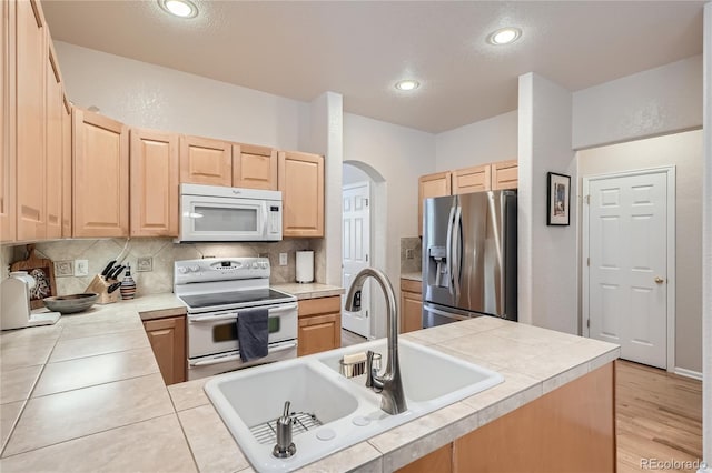 kitchen featuring tile counters, light brown cabinets, a sink, white appliances, and a peninsula