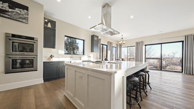 kitchen with stainless steel double oven, wood-type flooring, island exhaust hood, a kitchen island with sink, and gray cabinetry