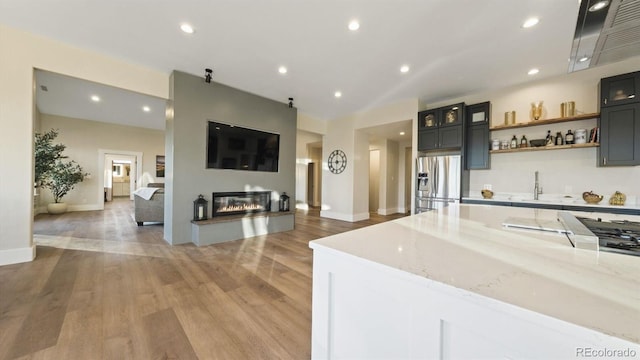 kitchen featuring light stone counters, stainless steel refrigerator with ice dispenser, light wood finished floors, recessed lighting, and a glass covered fireplace