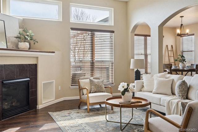 living room with baseboards, arched walkways, a tiled fireplace, dark wood-style flooring, and a notable chandelier