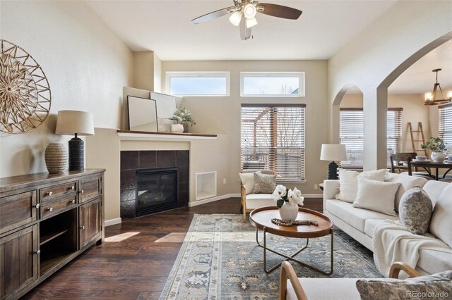 living area featuring dark wood-style floors, arched walkways, a fireplace, baseboards, and ceiling fan with notable chandelier