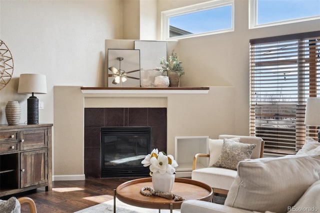 living area featuring baseboards, dark wood finished floors, a tiled fireplace, and ceiling fan