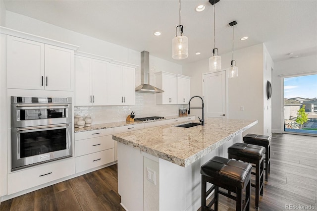 7169 Canyon Sky Trl, Castle Pines, CO 80108 - kitchen featuring wall chimney range hood, a sink, stainless steel double oven, dark wood-type flooring, and a kitchen island with sink