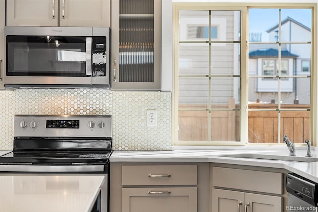 kitchen with gray cabinetry, a sink, light countertops, appliances with stainless steel finishes, and glass insert cabinets