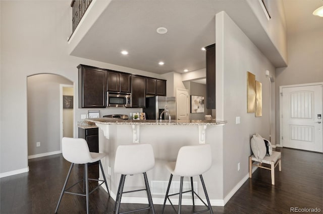 kitchen featuring appliances with stainless steel finishes, a kitchen bar, dark brown cabinets, arched walkways, and light stone counters