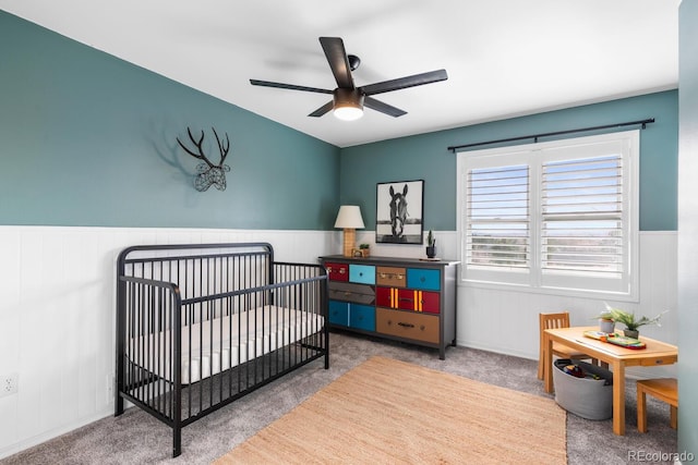 carpeted bedroom featuring a crib, a ceiling fan, and a wainscoted wall