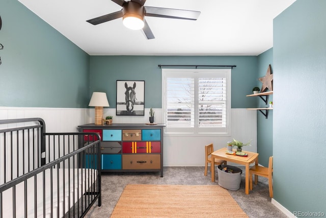 carpeted bedroom featuring wainscoting, a crib, and ceiling fan