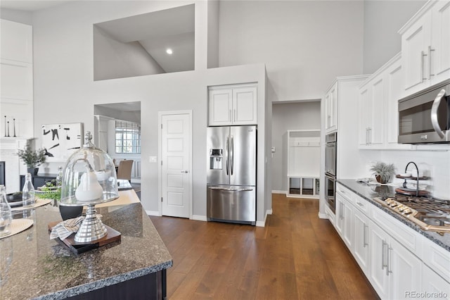 kitchen featuring dark stone counters, a towering ceiling, dark wood-style floors, appliances with stainless steel finishes, and white cabinetry