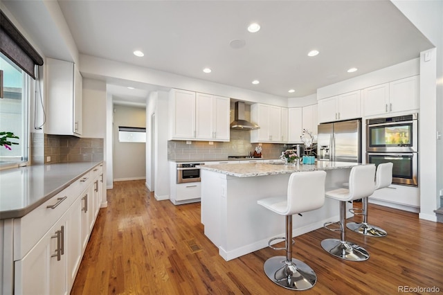 kitchen with an island with sink, white cabinets, stainless steel appliances, and wall chimney range hood