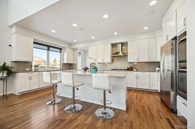 kitchen with a breakfast bar, stainless steel appliances, wall chimney range hood, a center island, and white cabinetry