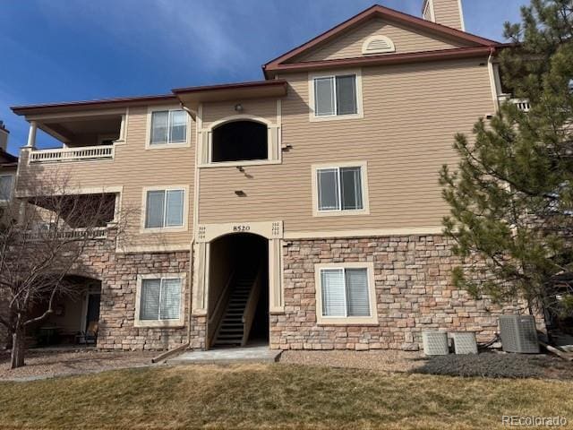 back of property with central AC unit, a lawn, a balcony, stone siding, and a chimney