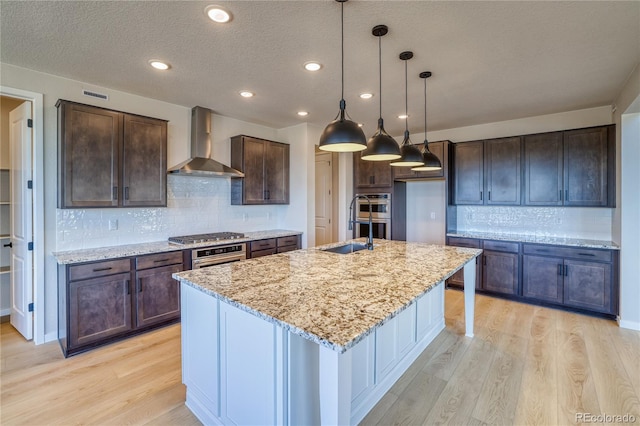 kitchen with light stone countertops, light wood-style floors, wall chimney range hood, an island with sink, and dark brown cabinets