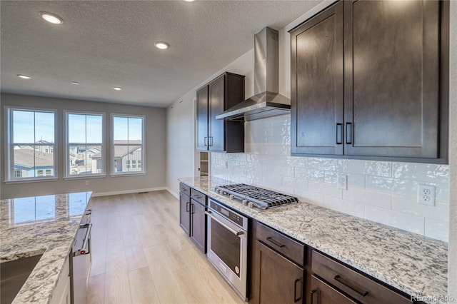 kitchen featuring light stone countertops, wall chimney exhaust hood, appliances with stainless steel finishes, and light wood-style flooring