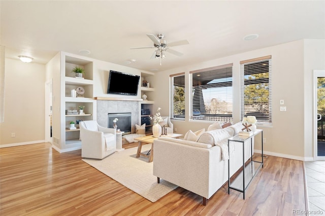 living room featuring a tiled fireplace, built in features, ceiling fan, and light hardwood / wood-style flooring