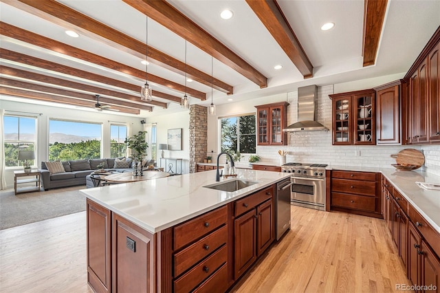 kitchen featuring wall chimney range hood, sink, stainless steel appliances, an island with sink, and decorative light fixtures