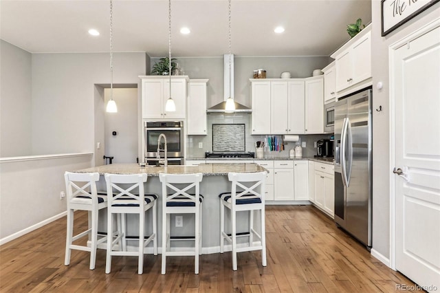 kitchen featuring white cabinetry, decorative light fixtures, stainless steel appliances, a kitchen island with sink, and wall chimney range hood