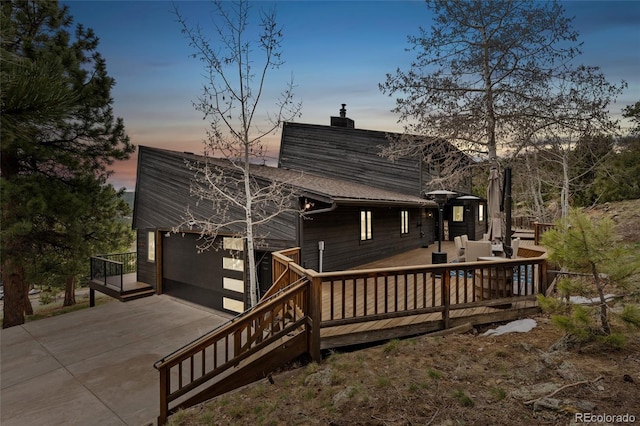 6640 Kilimanjaro Dr, Evergreen, CO 80439 - back of property at dusk with a garage, a wooden deck, roof with shingles, a chimney, and concrete driveway