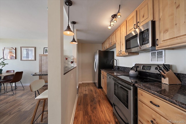 kitchen with light brown cabinets, dark stone counters, hanging light fixtures, appliances with stainless steel finishes, and dark hardwood / wood-style flooring