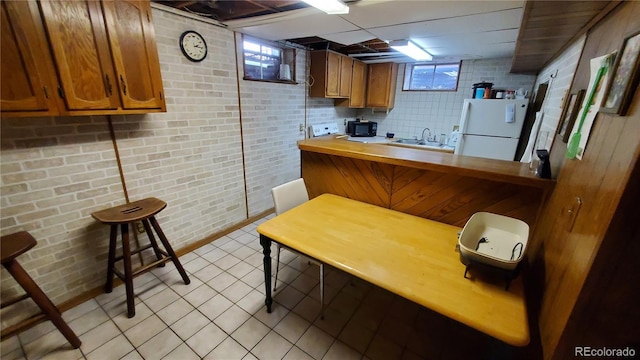 kitchen with brown cabinets, a sink, freestanding refrigerator, light tile patterned flooring, and brick wall