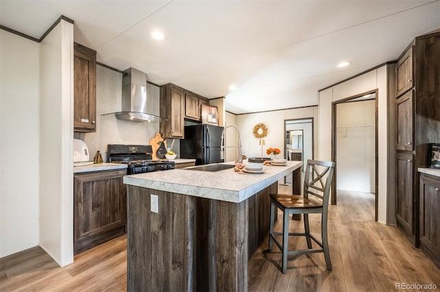 kitchen with a kitchen island with sink, light hardwood / wood-style floors, black appliances, and wall chimney range hood