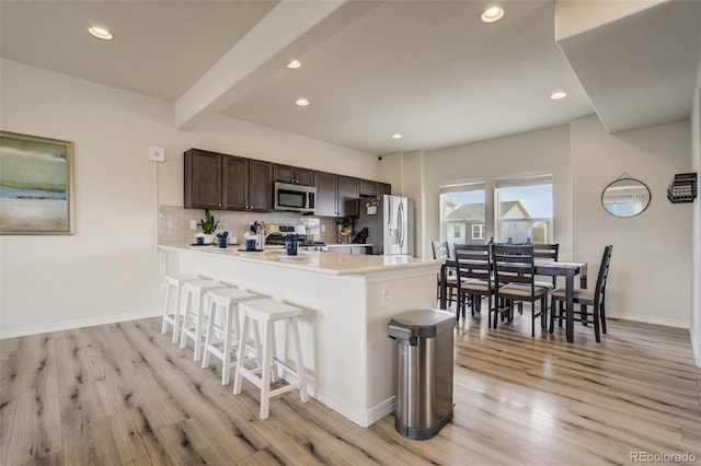 kitchen featuring a kitchen bar, backsplash, appliances with stainless steel finishes, light wood finished floors, and dark brown cabinets
