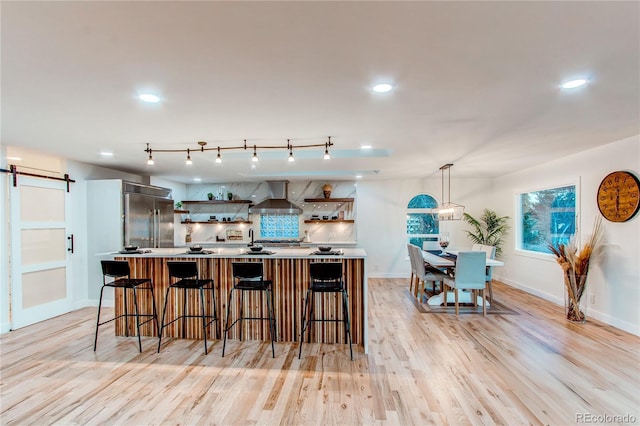 kitchen featuring a barn door, wall chimney range hood, light hardwood / wood-style floors, pendant lighting, and built in fridge