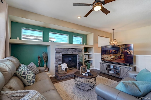 living room with built in shelves, ceiling fan with notable chandelier, a stone fireplace, and hardwood / wood-style flooring