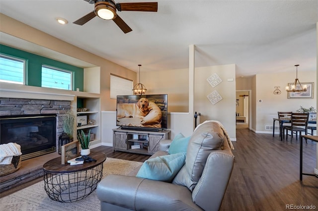 living room with built in shelves, dark hardwood / wood-style flooring, ceiling fan with notable chandelier, and a stone fireplace