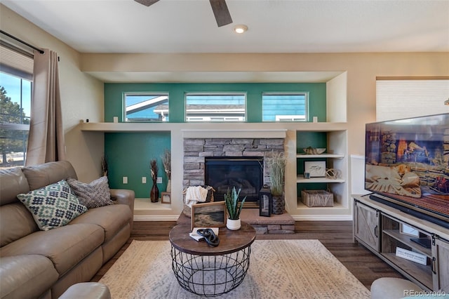 living room with ceiling fan, built in shelves, dark wood-type flooring, and a stone fireplace