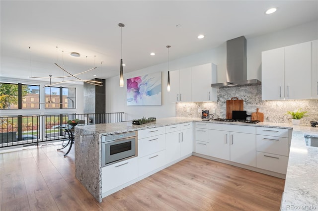 kitchen featuring hanging light fixtures, white cabinetry, light stone counters, a peninsula, and wall chimney exhaust hood