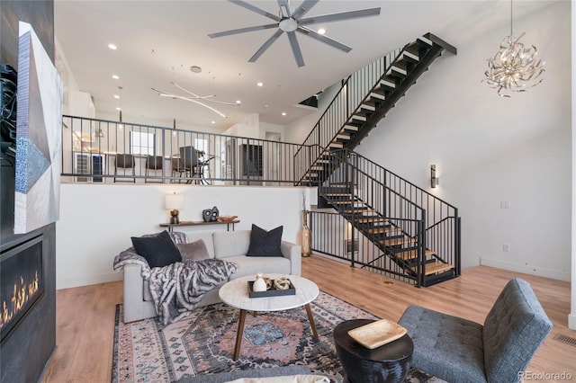 living area featuring stairs, wood finished floors, ceiling fan with notable chandelier, a glass covered fireplace, and recessed lighting