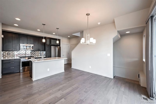 kitchen with hanging light fixtures, appliances with stainless steel finishes, light wood-type flooring, a center island with sink, and decorative backsplash