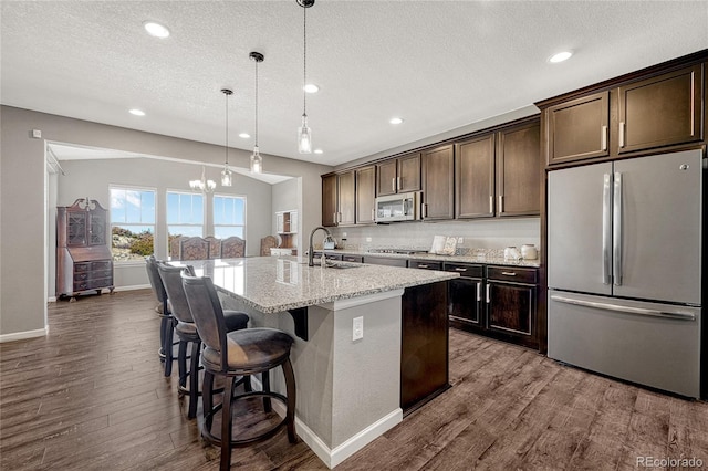 kitchen featuring dark brown cabinetry, light stone countertops, an island with sink, pendant lighting, and appliances with stainless steel finishes