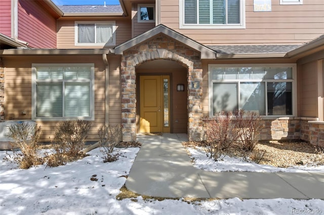 snow covered property entrance with stone siding and roof with shingles