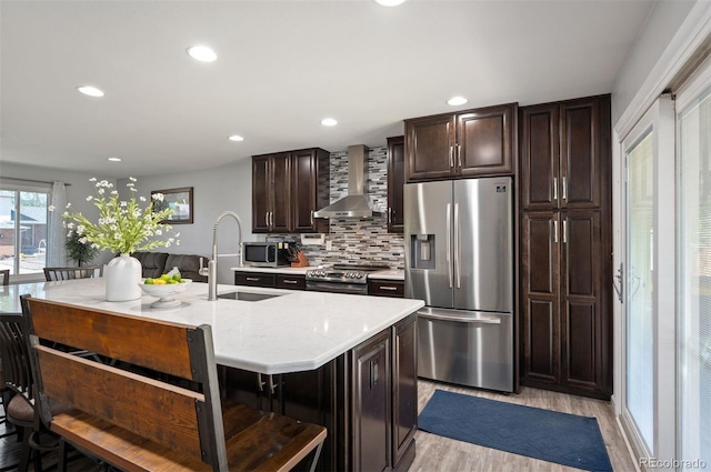 kitchen with stainless steel appliances, wall chimney range hood, a sink, light wood finished floors, and tasteful backsplash