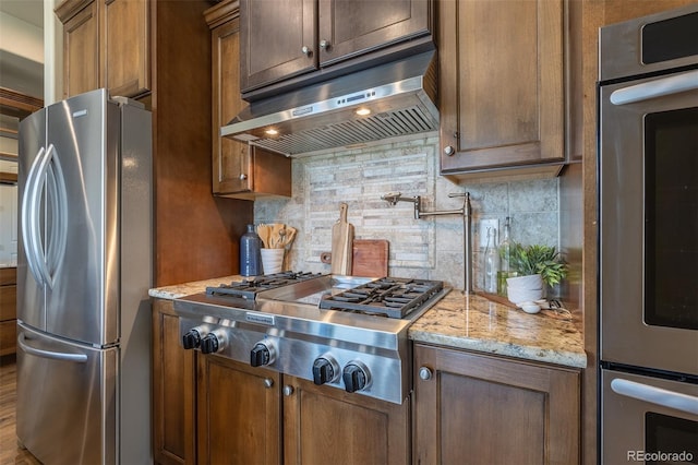 3012 Cathedral Park Vw, Colorado Springs, CO 80904 - kitchen with appliances with stainless steel finishes, under cabinet range hood, decorative backsplash, light stone countertops, and brown cabinets