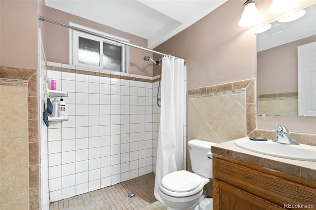 bathroom with a wainscoted wall, toilet, vanity, tiled shower, and tile walls