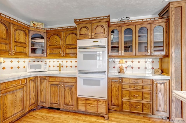 kitchen featuring white appliances, tile countertops, light wood finished floors, backsplash, and brown cabinetry