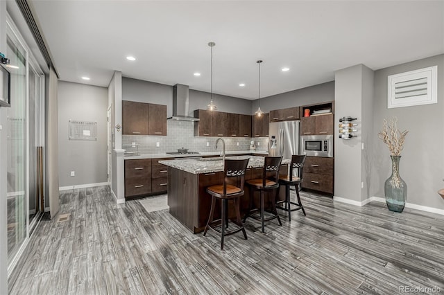 kitchen with a kitchen island with sink, stainless steel appliances, dark brown cabinetry, a kitchen breakfast bar, and wall chimney range hood