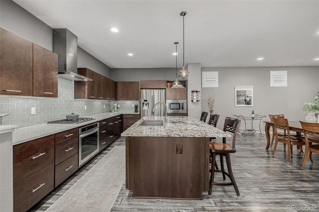 kitchen with a breakfast bar, a sink, appliances with stainless steel finishes, wall chimney range hood, and backsplash