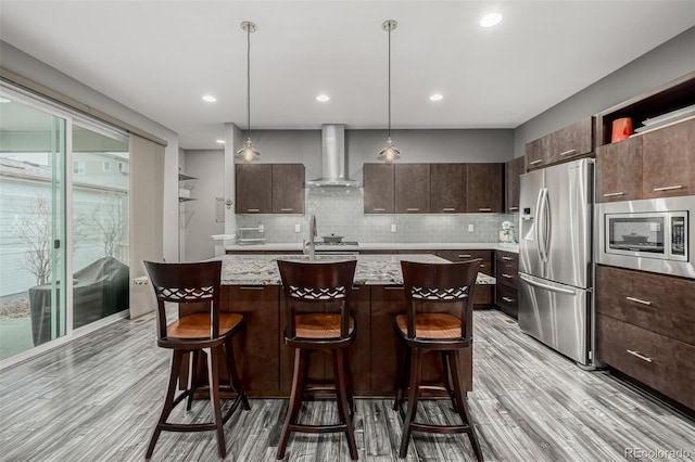 kitchen with dark brown cabinets, a breakfast bar area, an island with sink, appliances with stainless steel finishes, and wall chimney exhaust hood