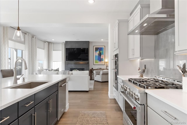 kitchen with stainless steel appliances, light countertops, light wood-style floors, a sink, and wall chimney range hood