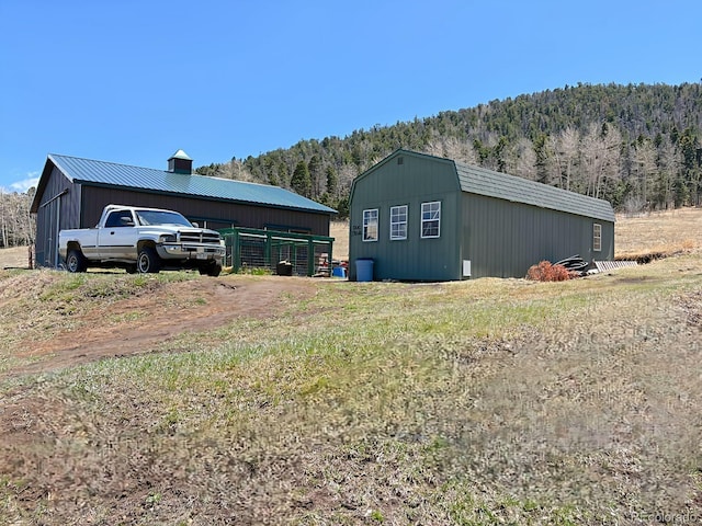 exterior space featuring a wooded view, a pole building, a chimney, metal roof, and an outbuilding