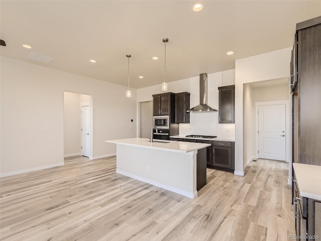 kitchen featuring a kitchen island with sink, wall chimney range hood, sink, hanging light fixtures, and stainless steel appliances