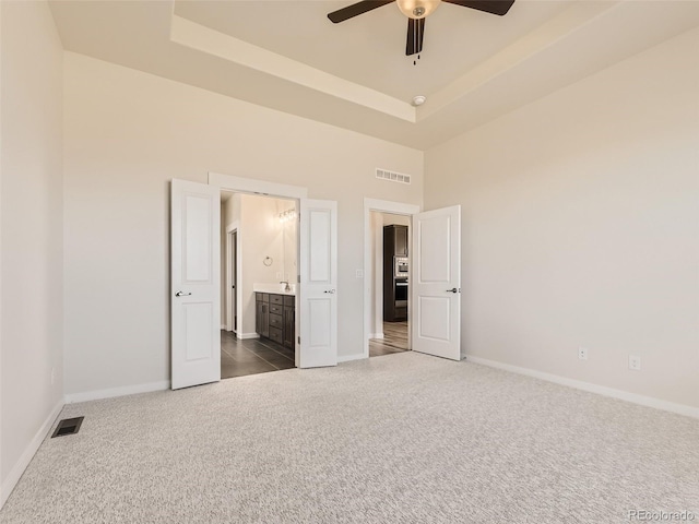 unfurnished bedroom with dark colored carpet, ensuite bathroom, ceiling fan, and a tray ceiling