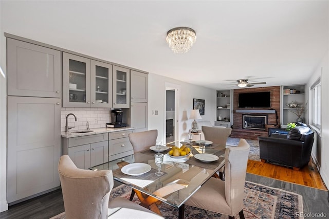 dining room with built in features, dark wood-type flooring, a brick fireplace, and ceiling fan with notable chandelier