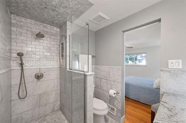 ensuite bathroom featuring toilet, a tile shower, ensuite bathroom, wood finished floors, and tile walls