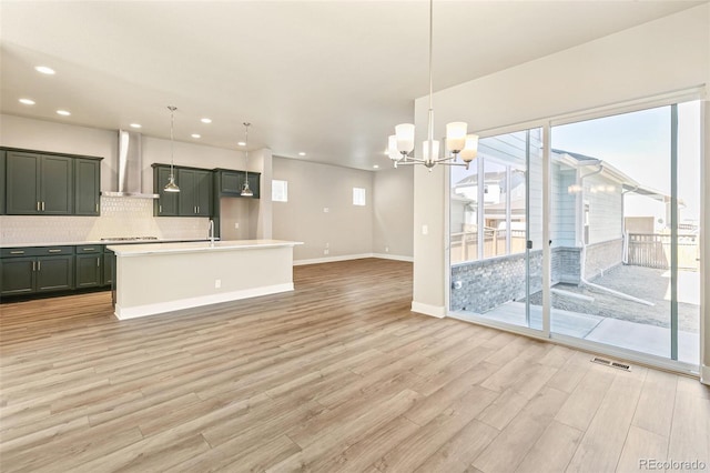 kitchen with pendant lighting, a center island with sink, wall chimney range hood, a notable chandelier, and light hardwood / wood-style floors