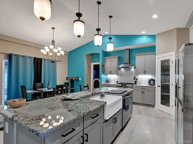 kitchen with gray cabinets, an island with sink, hanging light fixtures, and wall chimney range hood