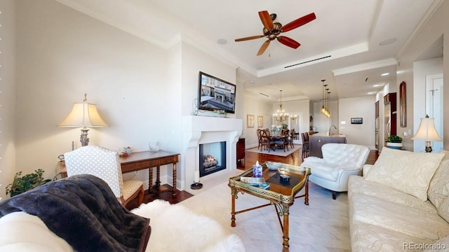 living area featuring a ceiling fan, a tray ceiling, crown molding, and a warm lit fireplace